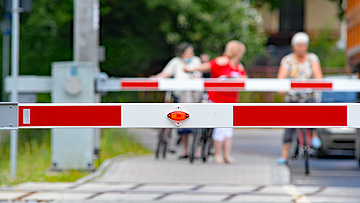 Zbliżenie na opuszczone rogatki, w tle trzy rowerzystki czekające przed przejazdem.