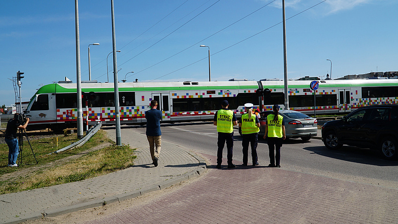 Przejazd kolejowy, przejeżdżający pociąg, przed przejazdem trzech funkcjonariuszy SOK, po prawej stronie samochody stojące przed przejazdem.