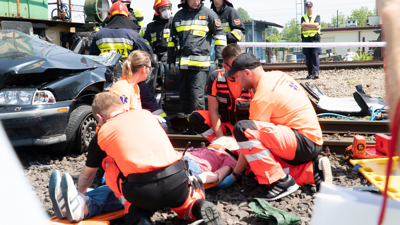 Czterech ratowników medycznych podczas udzielania pomocy osobie poszkodowanej. W tle strażacy i wrak samochodu.