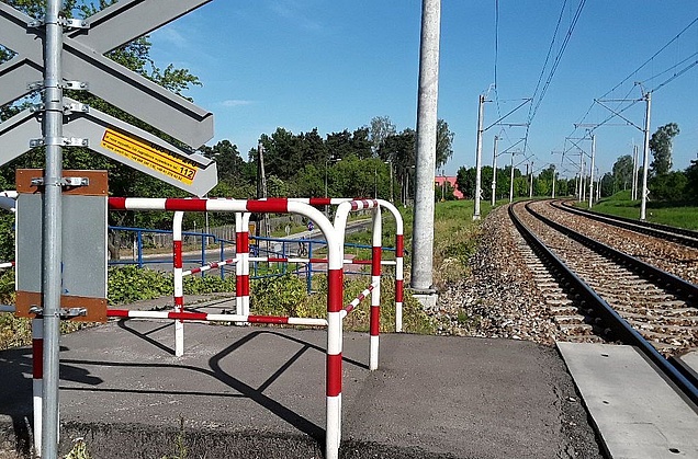 Przejście dla pieszych przy przejeździe kolejowym. Po lewej stronie tył znaku krzyża św. Andrzeja na którym znajduje się żółta naklejka PLK.