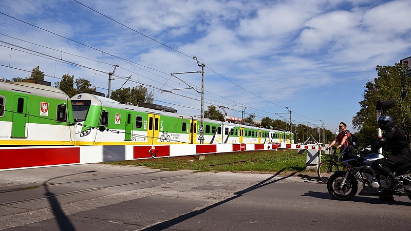 Rowerzysta i motocyklista stoją przed zamkniętym szlabanem. Pociąg przejeżdża przez przejazd